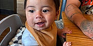 Bryson is registered to the contest to win money with this photo: toddler, child, baby, high_chair, blanket, bib, table, restaurant, wooden_table, person, indoor, smiling, happy, casual_clothing, tattoo, arm, seated, curly_hair, curious, closeup