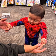 Bryson joined the competition — help win amazing prizes! toddler, child, costume, spiderman, standing, holding_hands, carpet, store, costume_shop, packaged_costumes, adult_hand, muscle_suit, red, blue, baby, indoor, shopping, floor, young_child, clothing