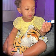 Aaliyah is registered to the contest to win money with this photo: toddler, child, yellow_dress, stuffed_toy, tiger, indoor, purple_lighting, marker, curious, sitting, toy, person, face, short_hair, cute, playful, room, soft_light, expression, holding