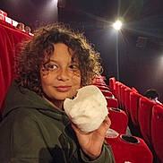 Tristan a rejoint le concours — aidez-le/la à gagner de superbes lots ! child, curly_hair, cotton_candy, movie_theater, red_seats, hoodie, snack, indoor, portrait, smile, lights, seat_cup_holder, casual_clothing, young_person, entertainment, leisure, fun, relaxed, people_in_background, seated
