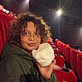 child, curly_hair, cotton_candy, movie_theater, red_seats, hoodie, snack, indoor, portrait, smile, lights, seat_cup_holder, casual_clothing, young_person, entertainment, leisure, fun, relaxed, people_in_background, seated