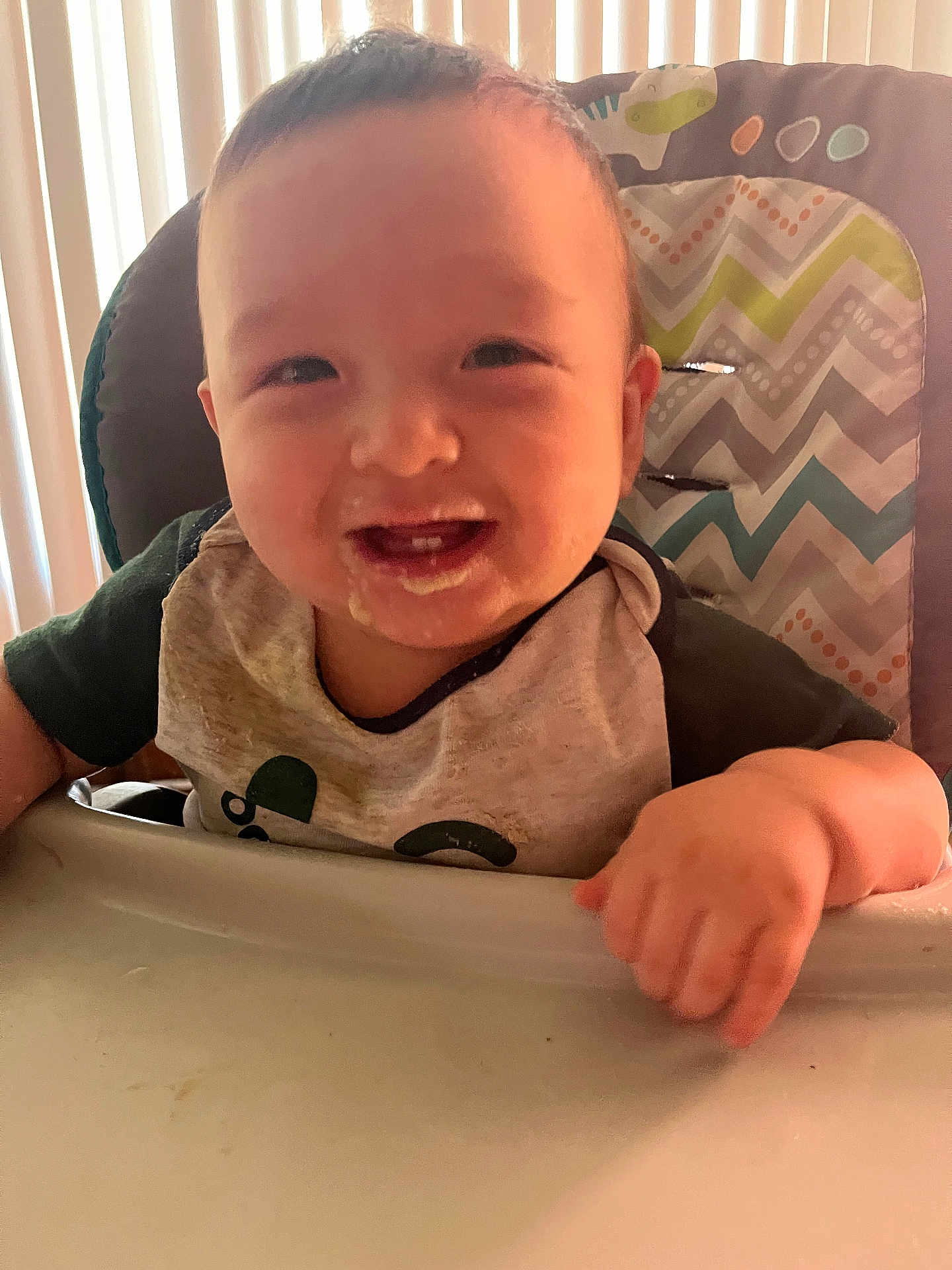 Martin is registered to the contest to win money with this photo: baby, high_chair, smiling, food, messy, bib, child, face, hand, teeth, indoor, light, window_blinds, clothing, person, cute, happy, infant, feeding, portrait