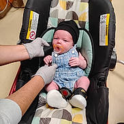 Liam is registered to the contest to win money with this photo: newborn, baby, infant, car_seat, pacifier, knit_cap, overalls, clothing, gloves, medical_exam, hospital, sitting, lying_down, portrait, face, indoors, colorful, safety_harness, shoes, caregiver
