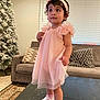 Tehreek is registered to the contest to win money with this photo: toddler, child, dress, headband, pink, shoes, socks, table, living_room, couch, cushion, christmas_tree, decor, window_blinds, floor, rug, person, indoor, curly_hair, cute