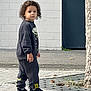 child, curly_hair, standing, sidewalk, urban, casual_clothing, sneakers, tree, pavement, curious, neutral_expression, outdoor, person, black_clothing, daylight, street, young, pants, sweatshirt, footwear