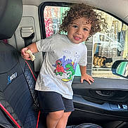 Ishaq a rejoint le concours — aidez-le/la à gagner de superbes lots ! child, toddler, curly_hair, barefoot, car_interior, car_seat, black_seat, bread, white_tshirt, shorts, smiling, window, door, seatbelt, shopping_display, clothing, hand, face, person, indoor