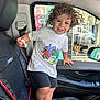 child, toddler, curly_hair, barefoot, car_interior, car_seat, black_seat, bread, white_tshirt, shorts, smiling, window, door, seatbelt, shopping_display, clothing, hand, face, person, indoor