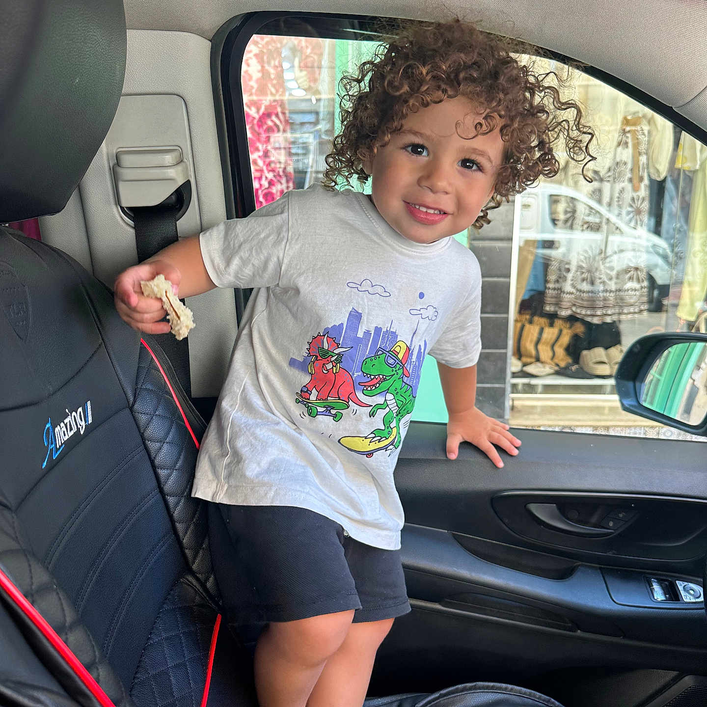 Ishaq a rejoint le concours — aidez-le/la à gagner de superbes lots ! barefoot, black_seat, bread, car_interior, car_seat, child, clothing, curly_hair, door, face, hand, indoor, person, seatbelt, shopping_display, shorts, smiling, toddler, white_tshirt, window