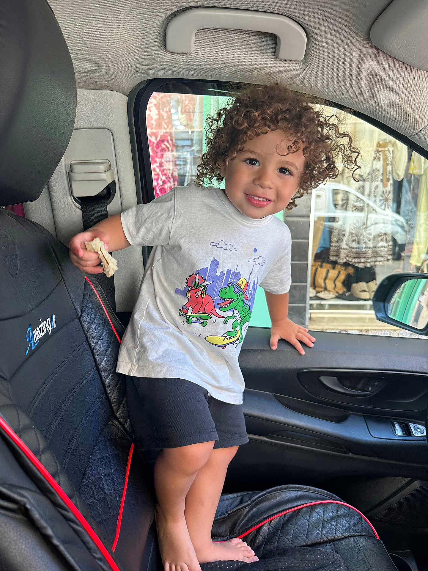 Ishaq a rejoint le concours — aidez-le/la à gagner de superbes lots ! child, toddler, curly_hair, barefoot, car_interior, car_seat, black_seat, bread, white_tshirt, shorts, smiling, window, door, seatbelt, shopping_display, clothing, hand, face, person, indoor