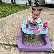 Mia is registered to the contest to win money with this photo: baby, child, walker, pigtails, smiling, outdoor, driveway, grass, car, stroller, chair, house, window, concrete, footwear, bib, toy, person, happy, curly_hair