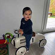 Timéo a rejoint le concours — aidez-le/la à gagner de superbes lots ! toddler, child, indoor, ride_on_toy, toy_car, flooring, window, natural_light, playmat, colorful_toys, jeans, sneakers, dark_sweater, smile, hair, person, steering_wheel, modern_home, light_wall, happy