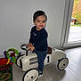 toddler, child, indoor, ride_on_toy, toy_car, flooring, window, natural_light, playmat, colorful_toys, jeans, sneakers, dark_sweater, smile, hair, person, steering_wheel, modern_home, light_wall, happy