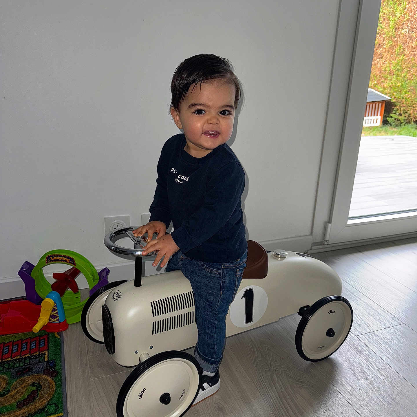 Timéo a rejoint le concours — aidez-le/la à gagner de superbes lots ! child, colorful_toys, dark_sweater, flooring, hair, happy, indoor, jeans, light_wall, modern_home, natural_light, person, playmat, ride_on_toy, smile, sneakers, steering_wheel, toddler, toy_car, window