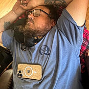 Peaches is registered to the contest to win money with this photo: man, dog, sleeping, tattoo, glasses, phone, blue_shirt, plaid_blanket, wooden_floor, beard, indoor, casual, resting, relaxed, pet, companion, close_up, chair, comfort, napping
