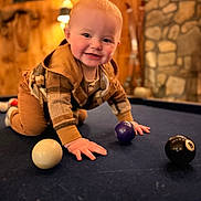 Jaxon is registered to the contest to win money with this photo: baby, child, crawling, pool_table, billiard_balls, indoor, wooden_wall, stone_wall, warm_lighting, smiling, cute, cozy, plaid_clothing, hands, face, floor, playing, person, infant, casual