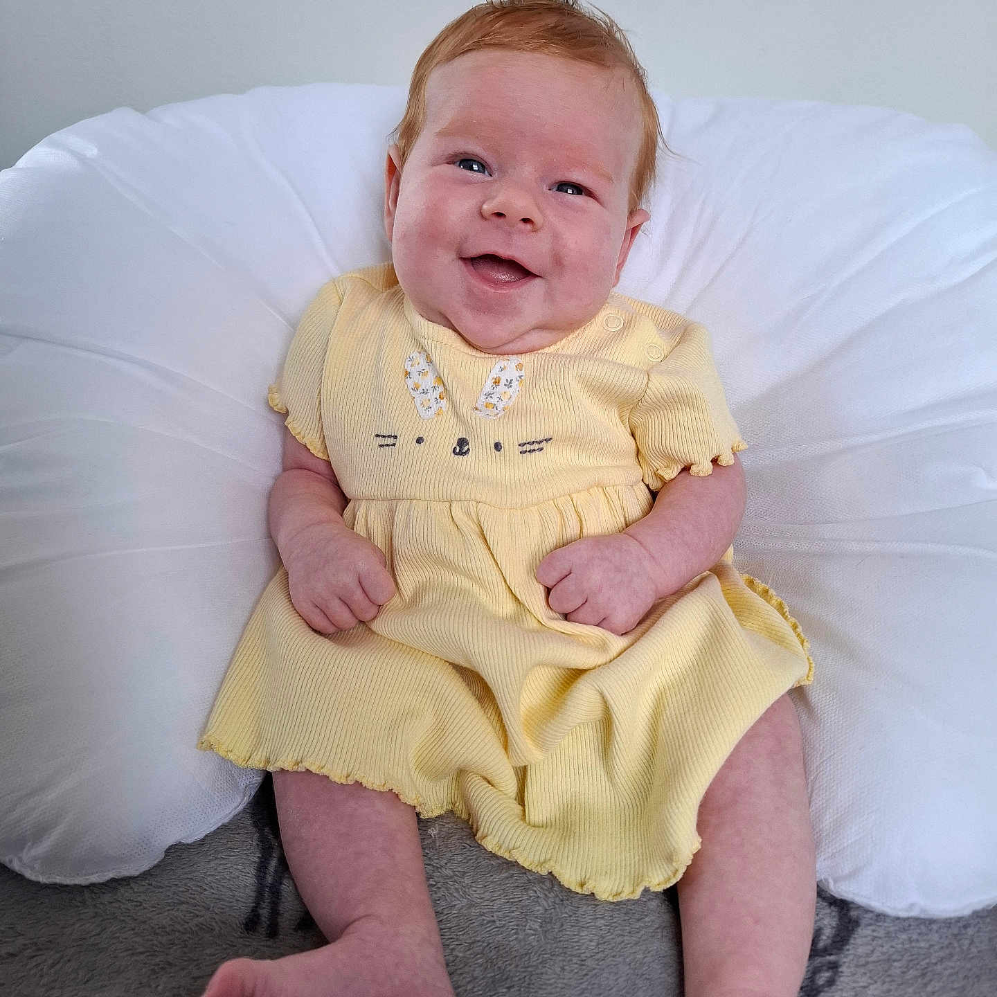 Poppy is registered to the contest to win money with this photo: baby, blanket, bunny_design, child, cushion, cute, face, feet, grey_blanket, hands, happy, indoors, infant, portrait, sitting, skin, smiling, soft_lighting, white_cushion, yellow_dress