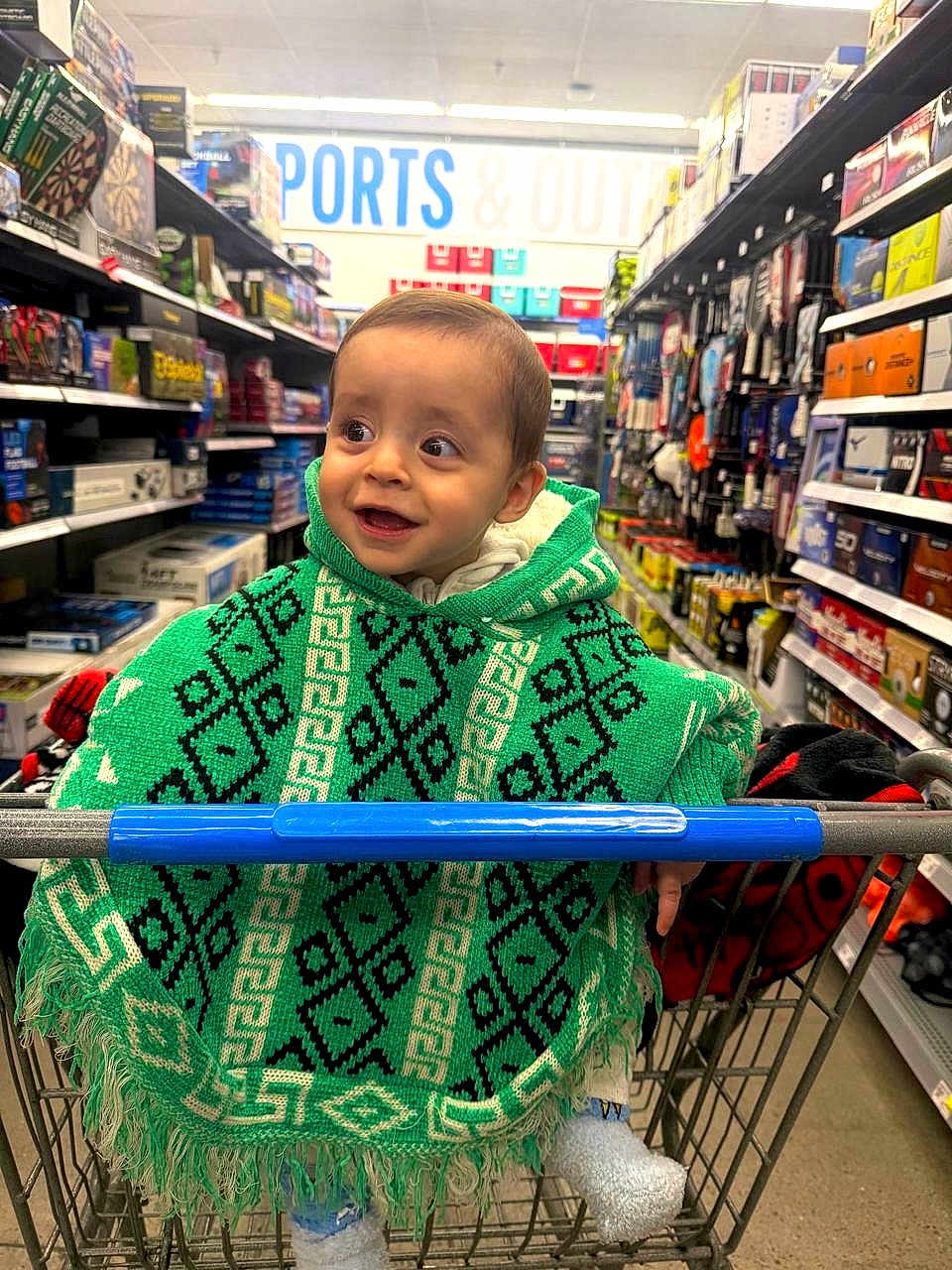 Winston joined the competition — help win amazing prizes! toddler, child, shopping_cart, green_poncho, store_aisle, toys, smiling, clothing, footwear, socks, indoor, retail, products, shelves, shopping, person, happy, cute, baby, portrait