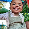 child, toddler, smiling, happy, playground, outdoor, portrait, face, teeth, dress, floral_shirt, overalls, braids, earrings, jungle_gym, slide, green_railing, blue_sky, daylight, playful