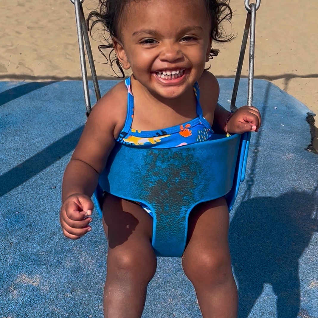 Maelynn joined the competition — help win amazing prizes! toddler, child, baby, smiling, happy, swing, playground, beach, sand, ocean, outdoor, summer, swimsuit, blue, curly_hair, portrait, sitting, play, sunlight, vacation
