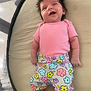 Delaney is registered to the contest to win money with this photo: baby, infant, smile, pink_shirt, colorful_pants, floral_pattern, lying_down, cushion, happy, cute, child, face, hair, indoors, playpen, newborn, clothing, person, skin, cheerful
