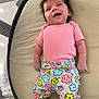 baby, infant, smile, pink_shirt, colorful_pants, floral_pattern, lying_down, cushion, happy, cute, child, face, hair, indoors, playpen, newborn, clothing, person, skin, cheerful