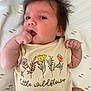 baby, infant, child, face, hand, clothing, shirt, text, floral_design, wild_hair, blanket, lying_down, cute, skin, portrait, young_child, indoors, soft_lighting, expression, small_fingers