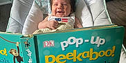 Delaney is registered to the contest to win money with this photo: baby, book, reading, pop_up_book, baby_chair, smiling, indoor, child, happy, hair, cushion, person, hand, floor, cabinet, wood_floor, furniture, colorful, toy, learning