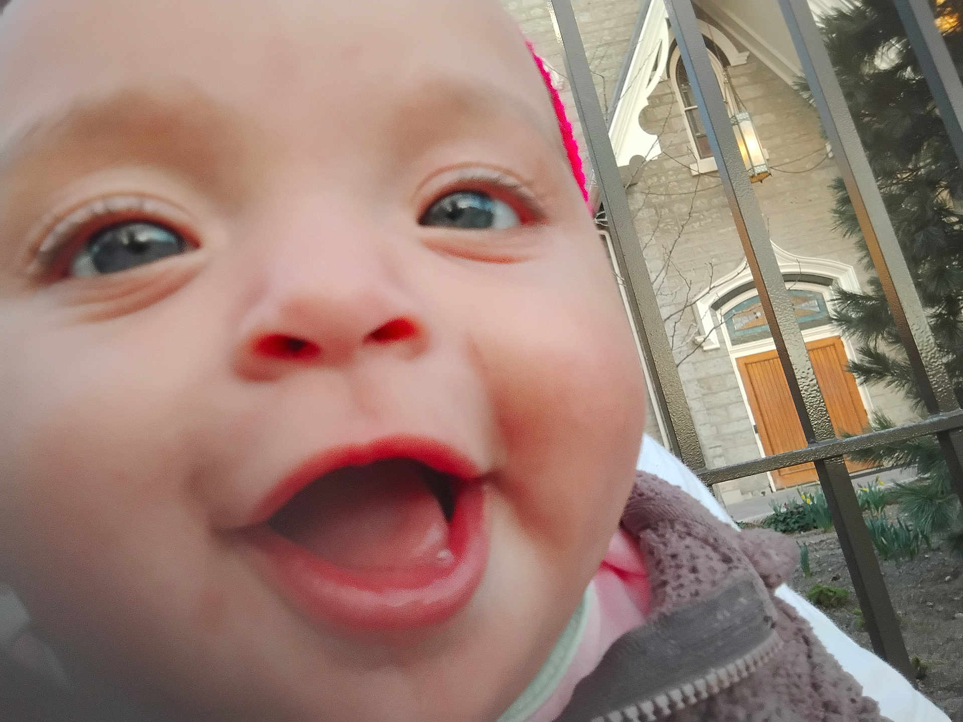 Miciah is registered to the contest to win money with this photo: baby, infant, child, close_up, face, smile, mouth_open, eyes, cheeks, outdoors, fence, railing, building, door, window, coat, hat, portrait, candid, giggling