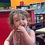 Haizel is registered to the contest to win money with this photo: toddler, child, curly_hair, purple_dress, high_chair, eating, crumbs, face, hand, indoor, arcade, colorful, red_wall, table, chair, person, portrait, messy, cute, snack