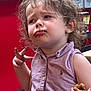 toddler, child, messy_face, purple_dress, curly_hair, food, chocolate, indoor, table, chair, expression, hand_gesture, snack, young_child, person, face, clothing, sitting, curious, brown_hair
