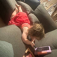 Haizel is registered to the contest to win money with this photo: toddler, child, red_dress, couch, tablet, toy, indoor, playing, curly_hair, pillow, fan, wooden_toy, floor, device, furniture, home, casual, relaxed, young_child, technology