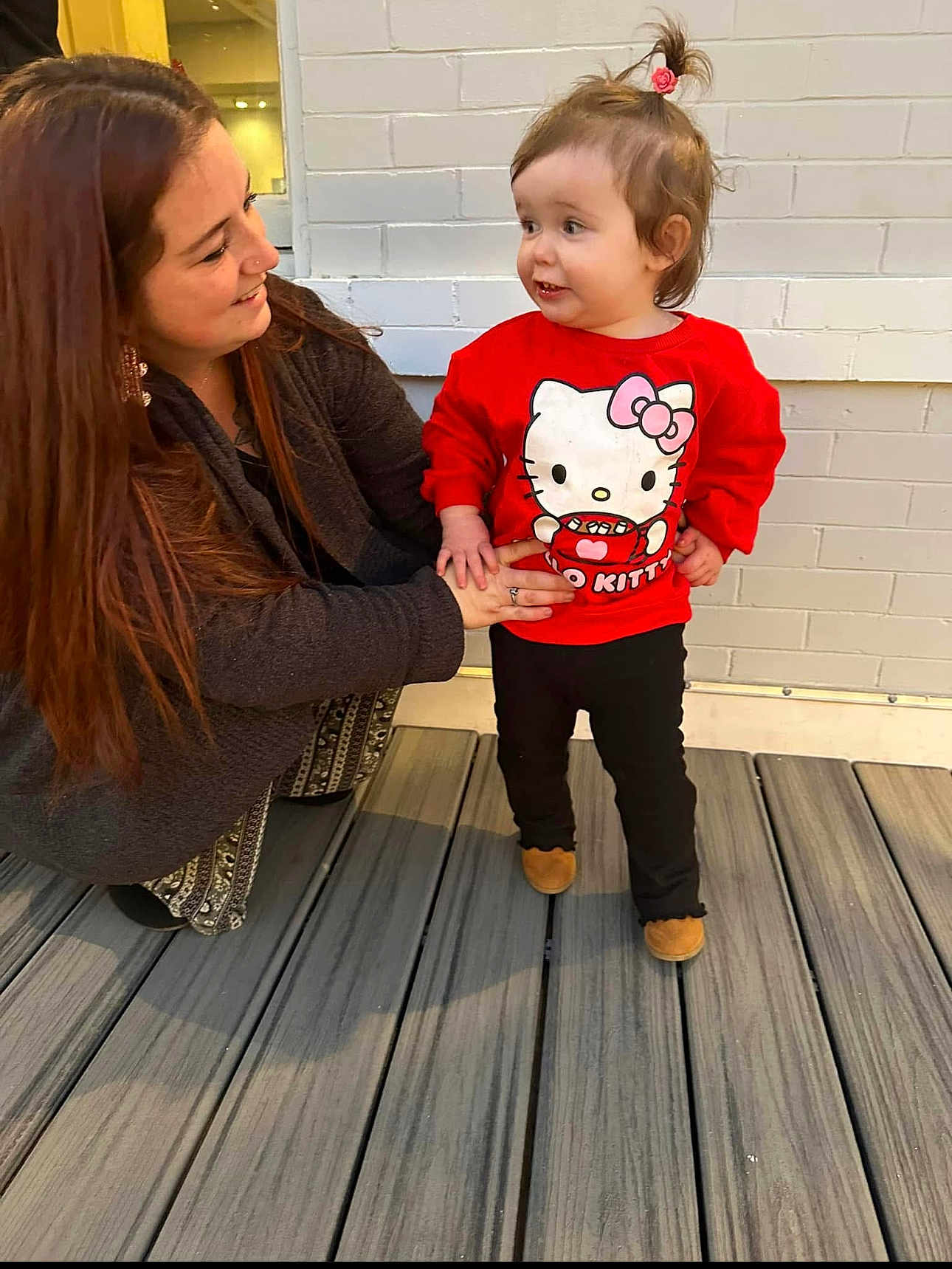 Haizel joined the competition — help win amazing prizes! toddler, woman, child, hello_kitty, red_sweatshirt, black_pants, brown_shoes, pony_tail, wooden_deck, brick_wall, smiling, holding_hands, outdoor, person, crouching, casual_clothing, affection, bonding, happy, daylight