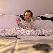 Sadon is registered to the contest to win money with this photo: toddler, child, bed, bedding, pillow, blanket, smile, happy, indoors, cozy, sleepwear, person, young_child, curly_hair, waving, soft_lighting, home, comfort, cute, portrait