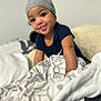 baby, child, hat, smile, bed, blanket, patterned_blanket, blue_shirt, indoor, cute, face, infant, person, cozy, young_child, playful, rest, comfort, portrait, headwear