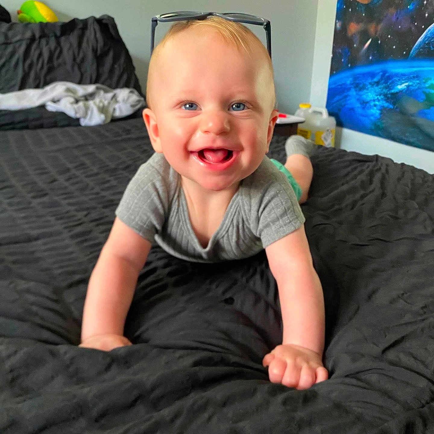Asher joined the competition — help win amazing prizes! baby, crawling, smiling, blue_eyes, gray_onesie, socks, bed, black_bedspread, sunglasses, indoor, poster, space_theme, wall_art, headwear, child, happy, person, room, cheerful, floor