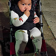 Matthew joined the competition — help win amazing prizes! baby, child, stroller, outdoor, sidewalk, night, portrait, sitting, face, hat, bear_hat, winter_clothing, black_vest, white_clothing, patterned_leggings, smiling, finger_in_mouth, street, pavement, casual