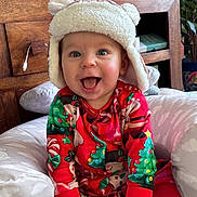 Archie is registered to the contest to win money with this photo: baby, child, smiling, hat, christmas_pajamas, holiday, indoor, cushion, wooden_furniture, plant, decorations, face, happy, sitting, toy, sock, red_clothing, cozy, cute, festive
