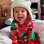 baby, child, smiling, hat, christmas_pajamas, holiday, indoor, cushion, wooden_furniture, plant, decorations, face, happy, sitting, toy, sock, red_clothing, cozy, cute, festive