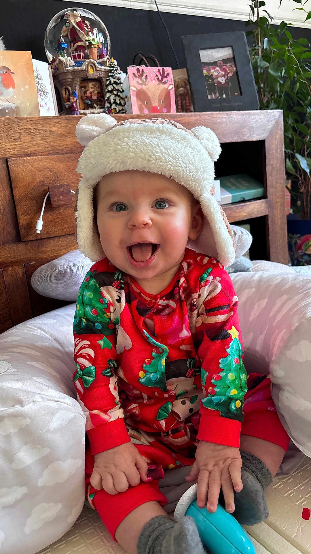 Archie is registered to the contest to win money with this photo: baby, child, smiling, hat, christmas_pajamas, holiday, indoor, cushion, wooden_furniture, plant, decorations, face, happy, sitting, toy, sock, red_clothing, cozy, cute, festive