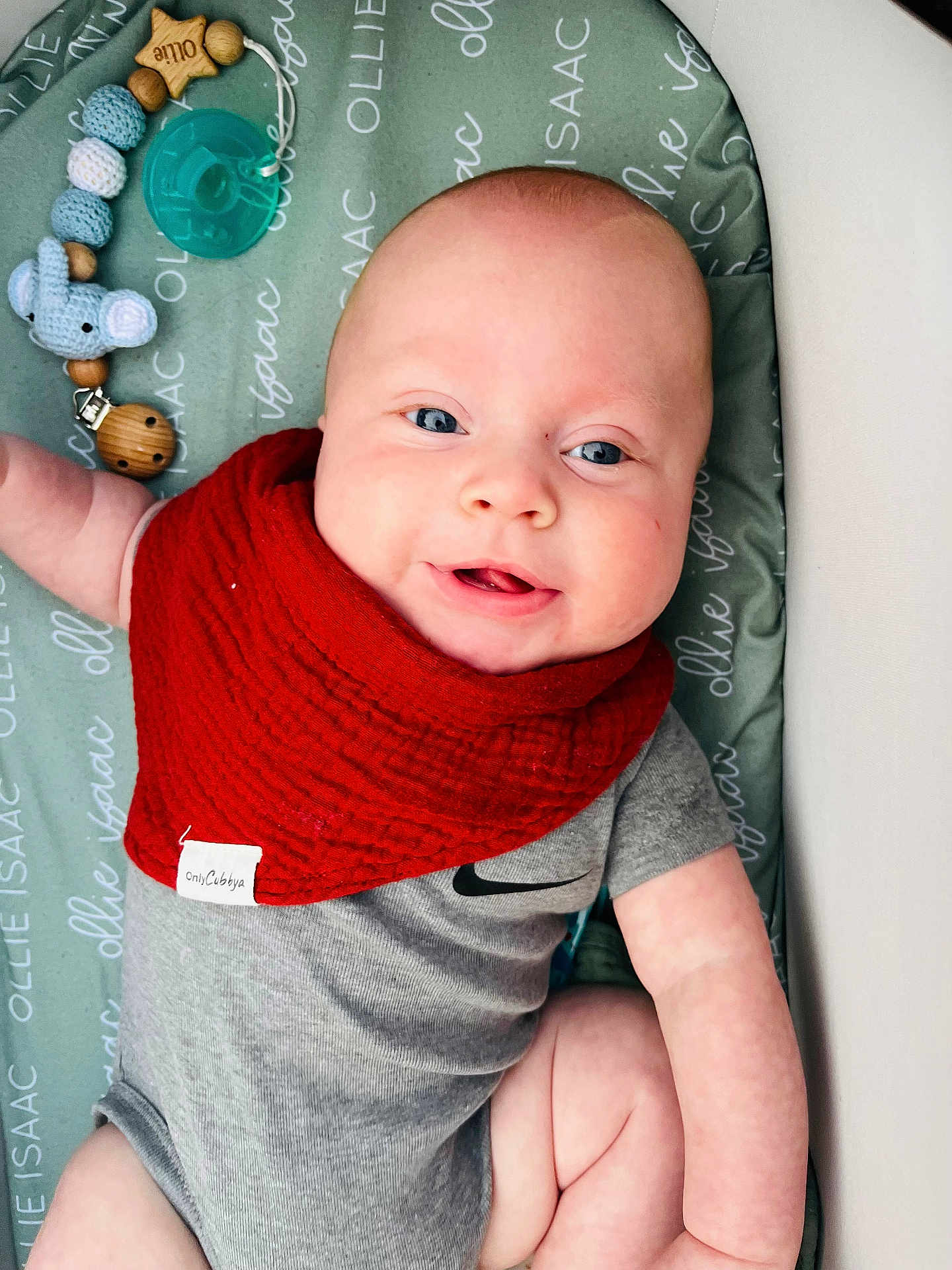 Ollie joined the competition — help win amazing prizes! baby, infant, smiling, red_bib, pacifier, toy_elephant, wooden_beads, grey_onesie, blue_eyes, chubby_cheeks, lying_down, changing_pad, patterned_text, close_up, portrait, skin, cute, nursery, accessory, arm