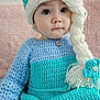 toddler, child, knitted_clothing, hat, braid, yarn, blue, turquoise, beads, earrings, sitting, portrait, indoor, soft_texture, pink_cushion, curly_hair_style, cute, young_child, face, eyes