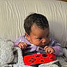 baby, child, controller, gaming, blanket, purple_onesie, couch, indoor, cozy, curious, face, hands, toy, soft_texture, relaxation, home, infant, person, expression, comfort