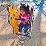 child, fence, handle, hedge, jeans, looking_sideways, outdoor, pink_sweater, playground, playtime, ponytails, rubber_surface, seat, shadow, sitting, sneakers, spring_ride, sunlight, toddler, yellow_play_equipment