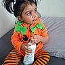 bed, big_eyes, bottle, child, curious_look, cute, dark_hair, face, hair_ties, hands, indoor, orange_jacket, pigtails, pillow, playtime, pumpkin_costume, sitting, striped_leggings, toddler, wall