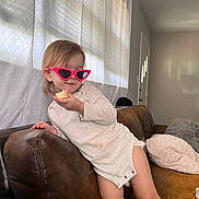 Annelise is registered to the contest to win money with this photo: toddler, child, sunglasses, pink, snack, couch, leather, living_room, curtains, window, casual_clothing, indoor, person, furniture, relaxed, cute, home, smile, hair, seat
