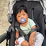 baby, stroller, pacifier, child, infant, outdoor, sidewalk, curious, black_stroller, casual_clothing, foot, holding, person, young_child, baby_clothes, daylight, urban, comfort, seated, transportation