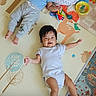 baby, infant, child, play_mat, toys, clothing, lying_down, floor, smiling, cute, indoors, colorful, person, portrait, happy, relaxed, sibling, home, casual, playtime