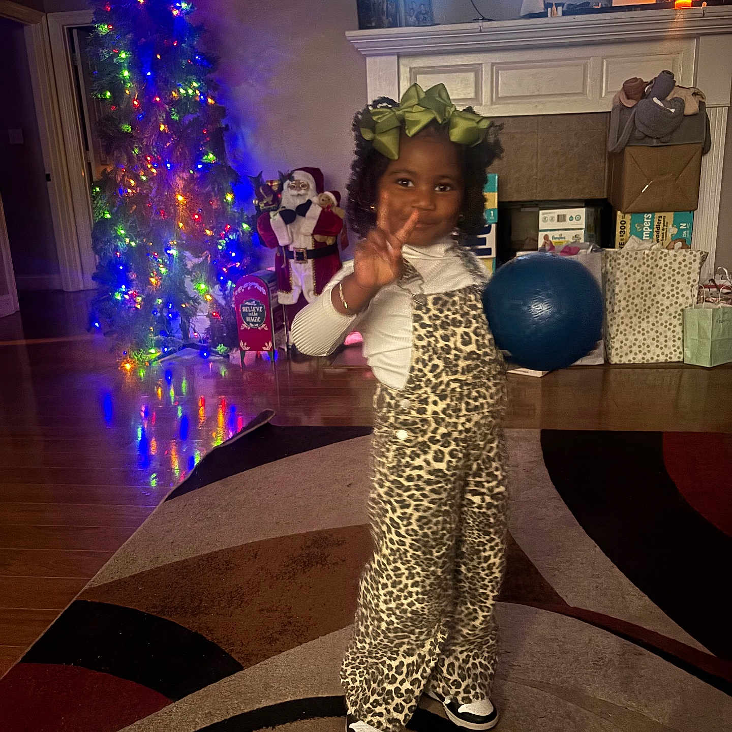 Demei joined the competition — help win amazing prizes! child, girl, leopard_print, overalls, green_bow, peace_sign, blue_ball, rug, living_room, christmas_tree, christmas_lights, gifts, holiday_decor, wooden_floor, fireplace, santa_figure, smiling, indoor, festive, happy