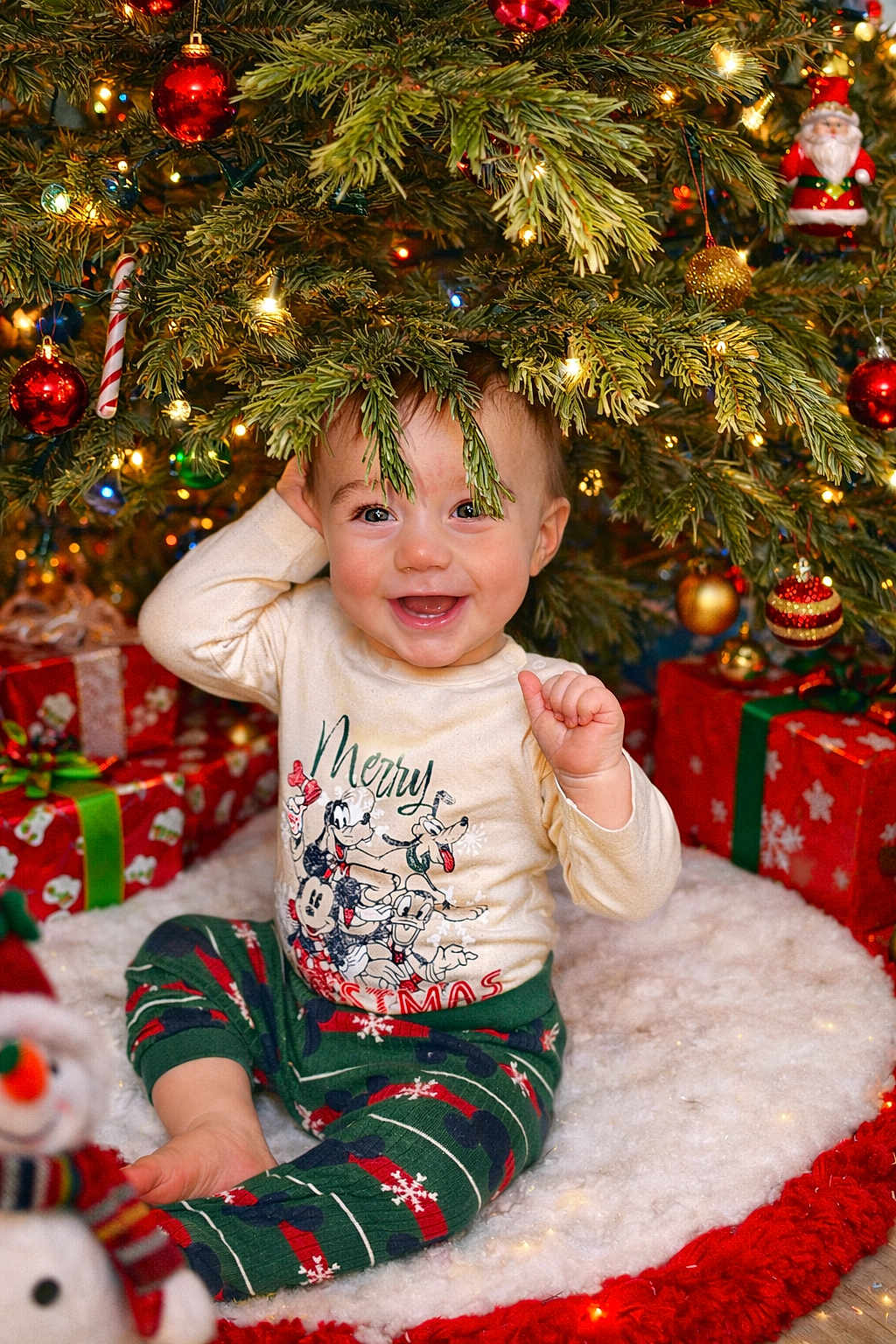 Jacob is registered to the contest to win money with this photo: baby, christmas_tree, christmas_ornaments, presents, holiday, festive_clothing, pajamas, smiling, indoors, holiday_lights, pine_branches, snowman_plush, child, celebration, happy, decorations, red, green, white, cute