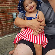 Azalea is registered to the contest to win money with this photo: toddler, child, smiling, hat, dress, stars, stripes, adult, lap, outside, brick_wall, concrete, person, happy, casual_clothing, summer, barefoot, family, candid, portrait